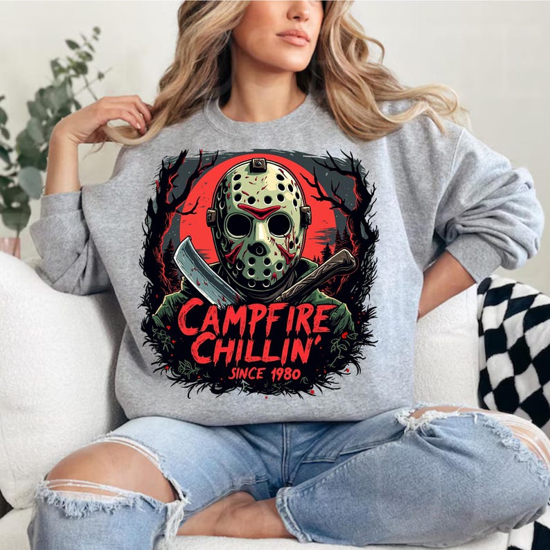 May include: A heather gray sweatshirt featuring a graphic of a hockey mask-wearing figure holding a machete and an axe. The design includes the text "CAMPFIRE CHILLIN' SINCE 1980" in red and white. The sweatshirt is a casual, long-sleeved garment.