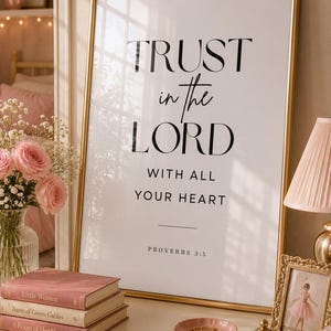 Puede incluir: Un cuadro enmarcado con el texto "TRUST in the LORD WITH ALL YOUR HEART" en escritura negra. El cuadro con marco dorado está sobre una cómoda blanca con libros, un joyero y una caja decorativa. También se ven rosas rosas y una lámpara.