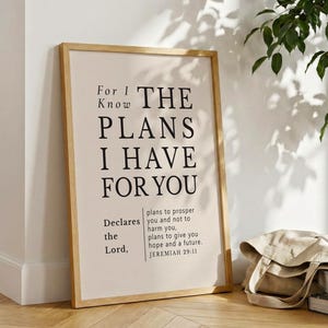 Puede incluir: Un cartel enmarcado con el texto "For I Know THE PLANS I HAVE FOR YOU" en fuente negra. El cartel está en un marco de madera clara y se apoya en una pared blanca. Una bolsa de lona y libros están en el suelo.