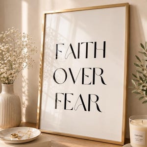 Puede incluir: Una impresión enmarcada con las palabras "FAITH OVER FEAR" en escritura negra. La obra de arte con marco dorado se exhibe sobre una superficie de madera, junto a un jarrón con flores, libros, una vela y un plato decorativo con joyas.