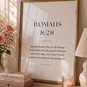 Könnte beinhalten: Goldgerahmter Druck mit dem Text „ROMANS 8:28“. Auf einer Holzoberfläche mit einer Blumenvase, Büchern, einer Kerze und einer Lampe.