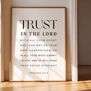 Può includere: Stampa incorniciata con le parole "TRUST IN THE LORD" in un carattere nero minimalista. Il testo continua con una citazione da Proverbi 3:5-6. La stampa è in una cornice di legno chiaro, appoggiata a un muro.