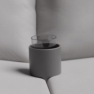 Puede incluir: Un portavasos gris con un vaso dentro, colocado en un sofá blanco. Una almohada gris está a la izquierda. El portavasos es cilíndrico y está diseñado para sostener un vaso o taza.