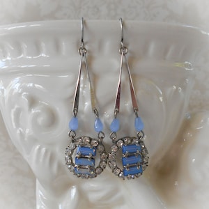 May include: A pair of drop earrings featuring silver-toned metal hooks and triangular frames. Each earring includes a small light blue bead above a circular pendant set with clear rhinestones surrounding three horizontal light blue rectangular stones.