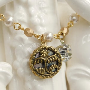 May include: A gold-toned necklace featuring a round, ornate pendant with floral detailing and a clear crystal charm. The necklace includes faux pearls and gold-colored beads, creating an elegant and vintage aesthetic.