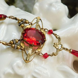 May include: An antique gold-toned necklace with large, faceted red gemstones and smaller red accents. The necklace features an ornate design with a central oval stone and a teardrop pendant. The jewelry is displayed on a white surface.