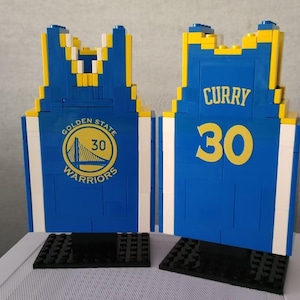 Peut inclure: Deux maillots de basket-ball bleus et jaunes en blocs de construction. L'un affiche le logo des Golden State Warriors et le numéro 30. L'autre affiche le nom "CURRY" et le numéro 30.