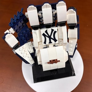 Op de afbeelding: Een Lego-sculptuur van een hand in wit, marineblauw en zwart. De hand houdt het logo van de New York Yankees vast. De sculptuur staat op een wit bord.