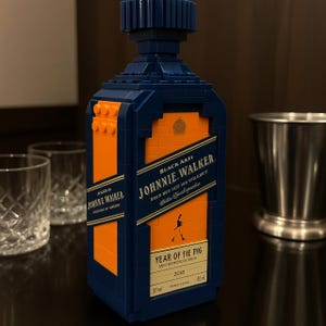 May include: A blue and orange block-built replica of a Johnnie Walker Black Label whiskey bottle. The label includes the text "Johnnie Walker" and "Year of the Pig 2018". Two clear glasses and a metal jigger are in the background.
