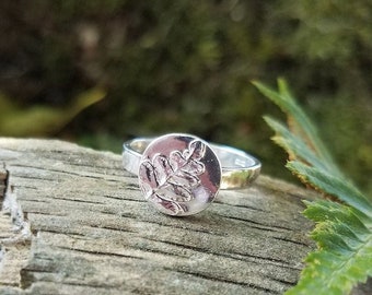 May include: A silver ring with a fern leaf design. The ring is on a piece of wood with a green leaf in the background.