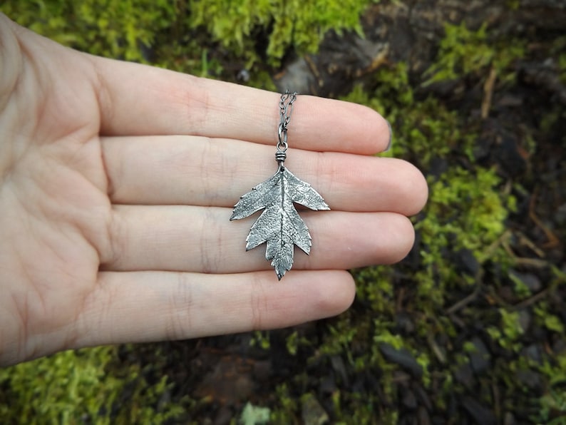 Puede incluir: Un collar con un colgante de hoja de plata en una cadena delicada. La hoja est&aacute; detallada con una superficie texturizada, lo que le da un aspecto realista.