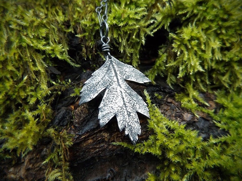 Puede incluir: Un collar con un colgante de hoja de plata, con una superficie texturizada detallada. El colgante est&aacute; colgado de una cadena de plata y se coloca sobre un fondo de musgo verde.