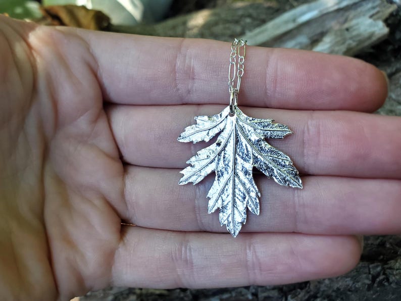 Puede incluir: Un collar de plata con un colgante de hoja texturizado y detallado. La hoja es una representaci&oacute;n realista de una hoja de arce.