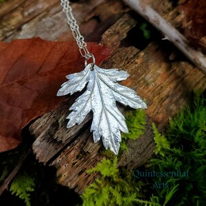 Puede incluir: Un collar de plata con un colgante de hoja detallada. El colgante est&aacute; hecho de metal y tiene una superficie texturizada. El collar est&aacute; fotografiado sobre un trozo de madera con musgo.