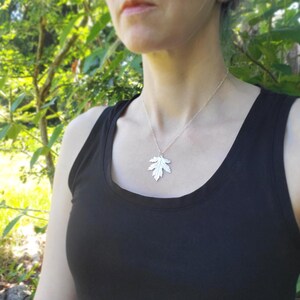 Puede incluir: Un collar de plata con un colgante de hoja detallada. El collar lo lleva una persona con una camiseta sin mangas negra.