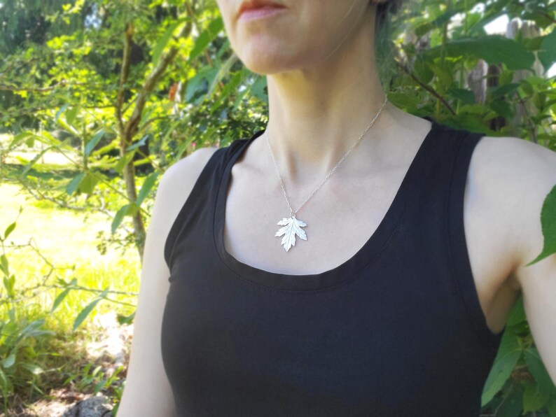 Puede incluir: Un collar de plata con un colgante de hoja detallada. El collar lo lleva una persona con una camiseta sin mangas negra.