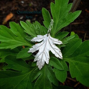 Puede incluir: Un collar con un colgante de hoja de plata en una cadena delicada. El colgante es una r&eacute;plica detallada de una hoja real, con venas y bordes claramente visibles. El collar se muestra sobre una cama de hojas verdes.