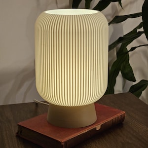 Peut inclure: Une lampe de table crème, côtelée, avec un abat-jour cylindrique et une base ronde. La lampe est allumée, posée sur un livre vintage à couverture rouge. Le cordon de la lampe est visible.