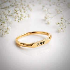 Peut inclure: Une bague en or poli avec une surface frontale plate et rectangulaire gravée des lettres M et K séparées par un petit symbole de cœur. La bague présente une finition métallique lisse et réfléchissante ainsi qu'un design circulaire simple.