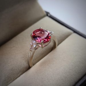 Puede incluir: Anillo de plata con una gran gema rosa de talla ovalada como pieza central, flanqueada por dos piedras claras más pequeñas en forma de pera. El anillo está engastado en un engaste clásico de garras.