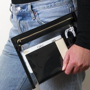 May include: A clear, rectangular clutch with a black leather trim and a gold zipper. Inside, a black wallet, a cream-colored card holder, and a white cable are visible. The word "FIRST" is printed on a white card.