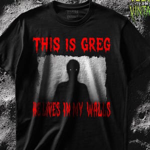 This horror T-shirt features a shadowy, humanoid figure with glowing eyes standing behind a textured, distorted wall pattern. Above it, the phrase “THIS IS GREG” is written in bold, dripping red letters, with “HE LIVES IN MY WALLS” printed below in the same eerie style. The entire design has an analog horror texture, grainy shadows, and unsettling contrast, making it perfect for fans of creepy artwork, scary graphic tees, and surreal dark aesthetics.