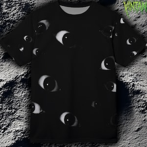 May include: Black t-shirt with a repeating pattern of white and grey eyes. The shirt has short sleeves and a crew neck. The text "VELOURA VINTANIQUE" is in green, stylized font on the upper right.