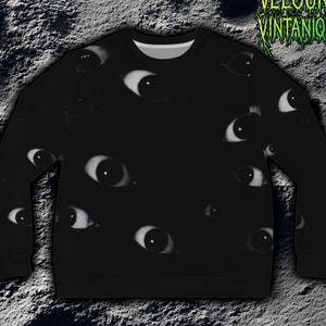 Könnte beinhalten: Schwarzes Sweatshirt mit einem sich wiederholenden Muster aus weißen und grauen Augen. Die Augen sind unterschiedlich groß und über die Vorderseite und die Ärmel verteilt. Der Text "VELOURA VINTANIQUE" ist in grüner, stilisierter Schrift.
