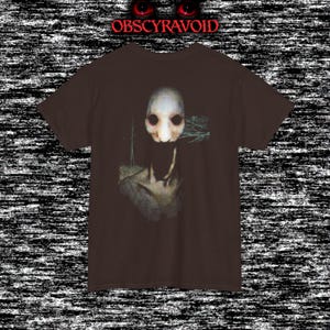 This analog horror T-shirt features a distressed VHS-style forest scene with eerie text overlay, combined with a terrifying pale mask-like creature emerging from darkness. Grainy textures, glitch effects, and cold blue tones create a creepy lost-footage atmosphere. Printed on a soft, high-quality shirt perfect for horror fans, dark aesthetic lovers, and anyone who enjoys unsettling, cinematic artwork.
