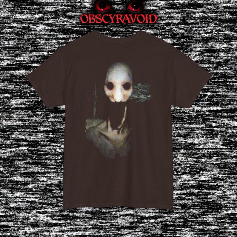 This analog horror T-shirt features a distressed VHS-style forest scene with eerie text overlay, combined with a terrifying pale mask-like creature emerging from darkness. Grainy textures, glitch effects, and cold blue tones create a creepy lost-footage atmosphere. Printed on a soft, high-quality shirt perfect for horror fans, dark aesthetic lovers, and anyone who enjoys unsettling, cinematic artwork.