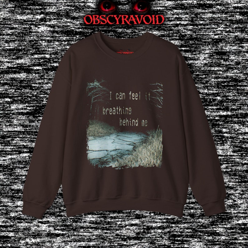 This analog horror sweatshirt features a chilling dual-sided design. The front shows a dark, grainy forest scene with glitchy text reading “I can feel it breathing behind me.” The back displays a disturbing pale creature emerging from the shadows, created with VHS static, noise, and eerie textures. Perfect for horror fans who love unsettling artwork, dark aesthetics, creepy forest themes, and lost-footage style designs.