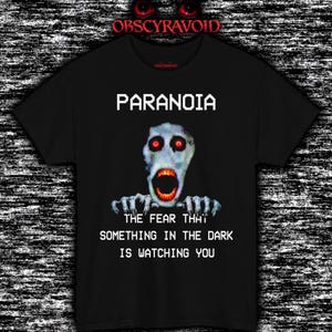 Può includere: Una t-shirt nera a maniche corte con la grafica di una creatura pallida dagli occhi sgranati, con occhi rossi luminosi e bocca aperta. Il testo PARANOIA e THE FEAR THAT SOMETHING IN THE DARK IS WATCHING YOU è scritto in un font pixelato bianco.