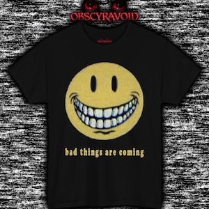 May include: Black t-shirt featuring a large yellow smiley face with a wide, unsettling grin. The text "bad things are coming" is printed below the face in a gold color. The shirt has the text "OBSCYRAVOID" at the top.