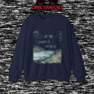 This analog horror sweatshirt features a chilling dual-sided design. The front shows a dark, grainy forest scene with glitchy text reading “I can feel it breathing behind me.” The back displays a disturbing pale creature emerging from the shadows, created with VHS static, noise, and eerie textures. Perfect for horror fans who love unsettling artwork, dark aesthetics, creepy forest themes, and lost-footage style designs.
