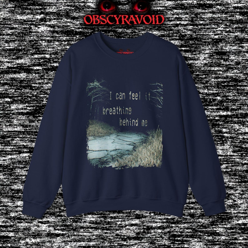 This analog horror sweatshirt features a chilling dual-sided design. The front shows a dark, grainy forest scene with glitchy text reading “I can feel it breathing behind me.” The back displays a disturbing pale creature emerging from the shadows, created with VHS static, noise, and eerie textures. Perfect for horror fans who love unsettling artwork, dark aesthetics, creepy forest themes, and lost-footage style designs.