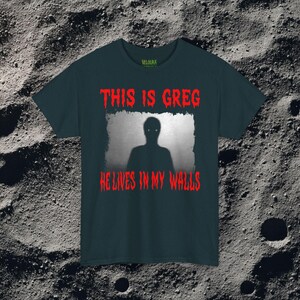 This horror T-shirt features a shadowy, humanoid figure with glowing eyes standing behind a textured, distorted wall pattern. Above it, the phrase “THIS IS GREG” is written in bold, dripping red letters, with “HE LIVES IN MY WALLS” printed below in the same eerie style. The entire design has an analog horror texture, grainy shadows, and unsettling contrast, making it perfect for fans of creepy artwork, scary graphic tees, and surreal dark aesthetics.