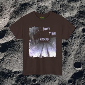 This analog horror-inspired T-shirt features the phrase “DON’T TURN AROUND” printed across a dark, eerie forest road under moonlight. On the other side, a creepy, distorted bunny character stares with a wide unsettling grin. The design blends glitchcore, VHS static, and nightmare-style textures for a perfect horror aesthetic. Printed on soft, high-quality fabric, ideal for horror fans and lovers of dark surreal art.
