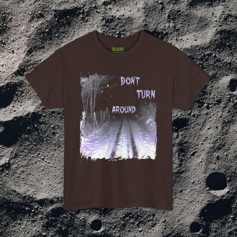 This analog horror-inspired T-shirt features the phrase “DON’T TURN AROUND” printed across a dark, eerie forest road under moonlight. On the other side, a creepy, distorted bunny character stares with a wide unsettling grin. The design blends glitchcore, VHS static, and nightmare-style textures for a perfect horror aesthetic. Printed on soft, high-quality fabric, ideal for horror fans and lovers of dark surreal art.