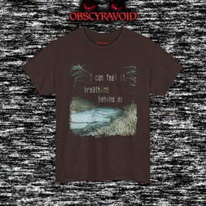 This analog horror T-shirt features a distressed VHS-style forest scene with eerie text overlay, combined with a terrifying pale mask-like creature emerging from darkness. Grainy textures, glitch effects, and cold blue tones create a creepy lost-footage atmosphere. Printed on a soft, high-quality shirt perfect for horror fans, dark aesthetic lovers, and anyone who enjoys unsettling, cinematic artwork.