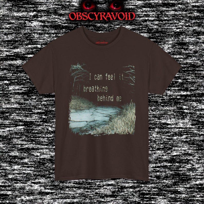 This analog horror T-shirt features a distressed VHS-style forest scene with eerie text overlay, combined with a terrifying pale mask-like creature emerging from darkness. Grainy textures, glitch effects, and cold blue tones create a creepy lost-footage atmosphere. Printed on a soft, high-quality shirt perfect for horror fans, dark aesthetic lovers, and anyone who enjoys unsettling, cinematic artwork.
