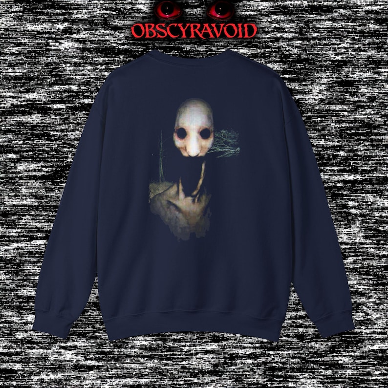This analog horror sweatshirt features a chilling dual-sided design. The front shows a dark, grainy forest scene with glitchy text reading “I can feel it breathing behind me.” The back displays a disturbing pale creature emerging from the shadows, created with VHS static, noise, and eerie textures. Perfect for horror fans who love unsettling artwork, dark aesthetics, creepy forest themes, and lost-footage style designs.