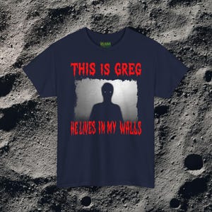 This horror T-shirt features a shadowy, humanoid figure with glowing eyes standing behind a textured, distorted wall pattern. Above it, the phrase “THIS IS GREG” is written in bold, dripping red letters, with “HE LIVES IN MY WALLS” printed below in the same eerie style. The entire design has an analog horror texture, grainy shadows, and unsettling contrast, making it perfect for fans of creepy artwork, scary graphic tees, and surreal dark aesthetics.