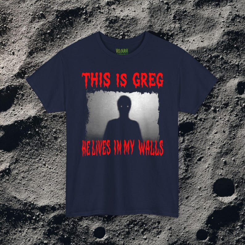 This horror T-shirt features a shadowy, humanoid figure with glowing eyes standing behind a textured, distorted wall pattern. Above it, the phrase “THIS IS GREG” is written in bold, dripping red letters, with “HE LIVES IN MY WALLS” printed below in the same eerie style. The entire design has an analog horror texture, grainy shadows, and unsettling contrast, making it perfect for fans of creepy artwork, scary graphic tees, and surreal dark aesthetics.