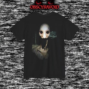 This analog horror T-shirt features a distressed VHS-style forest scene with eerie text overlay, combined with a terrifying pale mask-like creature emerging from darkness. Grainy textures, glitch effects, and cold blue tones create a creepy lost-footage atmosphere. Printed on a soft, high-quality shirt perfect for horror fans, dark aesthetic lovers, and anyone who enjoys unsettling, cinematic artwork.