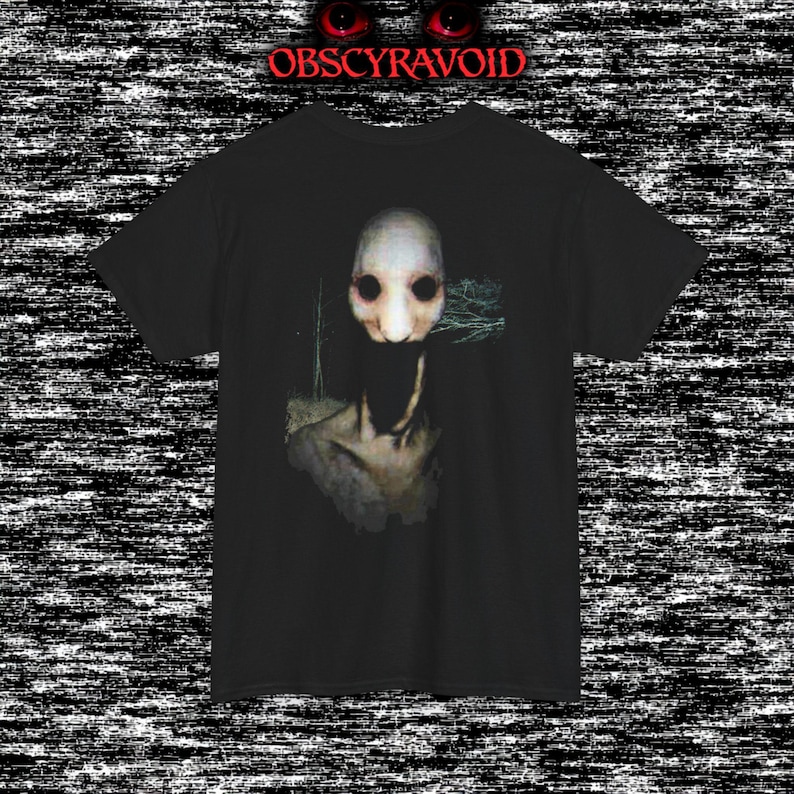 This analog horror T-shirt features a distressed VHS-style forest scene with eerie text overlay, combined with a terrifying pale mask-like creature emerging from darkness. Grainy textures, glitch effects, and cold blue tones create a creepy lost-footage atmosphere. Printed on a soft, high-quality shirt perfect for horror fans, dark aesthetic lovers, and anyone who enjoys unsettling, cinematic artwork.