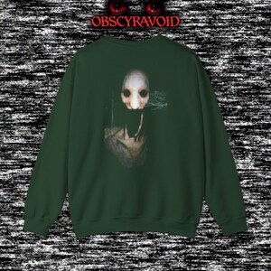 This analog horror sweatshirt features a chilling dual-sided design. The front shows a dark, grainy forest scene with glitchy text reading “I can feel it breathing behind me.” The back displays a disturbing pale creature emerging from the shadows, created with VHS static, noise, and eerie textures. Perfect for horror fans who love unsettling artwork, dark aesthetics, creepy forest themes, and lost-footage style designs.