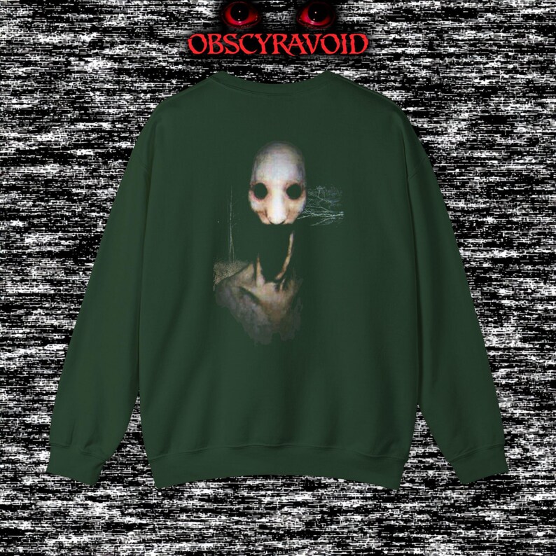 This analog horror sweatshirt features a chilling dual-sided design. The front shows a dark, grainy forest scene with glitchy text reading “I can feel it breathing behind me.” The back displays a disturbing pale creature emerging from the shadows, created with VHS static, noise, and eerie textures. Perfect for horror fans who love unsettling artwork, dark aesthetics, creepy forest themes, and lost-footage style designs.