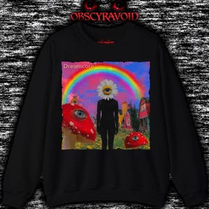 Könnte beinhalten: Ein schwarzes Sweatshirt mit einem Grafikdruck einer Figur mit Gänseblümchenkopf und einem Auge in der Mitte, die in einem Feld mit roten Pilzen mit Augen, einem Regenbogen am Himmel und Häusern steht, mit dem Text Dreamcore in der Ecke.