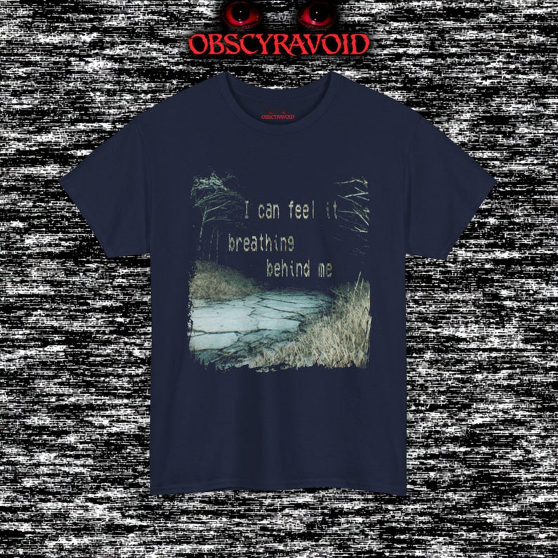 This analog horror T-shirt features a distressed VHS-style forest scene with eerie text overlay, combined with a terrifying pale mask-like creature emerging from darkness. Grainy textures, glitch effects, and cold blue tones create a creepy lost-footage atmosphere. Printed on a soft, high-quality shirt perfect for horror fans, dark aesthetic lovers, and anyone who enjoys unsettling, cinematic artwork.