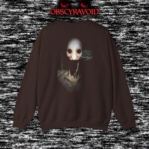 This analog horror sweatshirt features a chilling dual-sided design. The front shows a dark, grainy forest scene with glitchy text reading “I can feel it breathing behind me.” The back displays a disturbing pale creature emerging from the shadows, created with VHS static, noise, and eerie textures. Perfect for horror fans who love unsettling artwork, dark aesthetics, creepy forest themes, and lost-footage style designs.